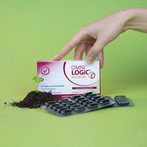 Eine Packung OMNi-LOGiC HUMIN mit Blistern und einem Häufchen Erde, aus dem eine Pflanze sprießt davor, wird von einer Frauenhand gehalten